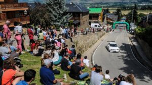 Gent asseguda a la gespa mentre mira un cotxe pujar per una carretera, amb les muntanyes de fons